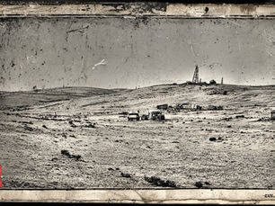 Old photographic representation of a cluster of buildings and vehicles in the desert near a radio tower.