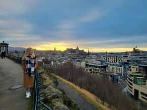 a blonde woman standing on a hill overlooking a city, Edinburgh. With sunset in the background.