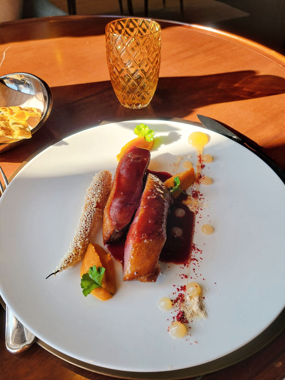 Gourmet dish on a white plate with duck, quinoa, sauce, and garnish. Amber glass in background. Sunlit wooden table setting.