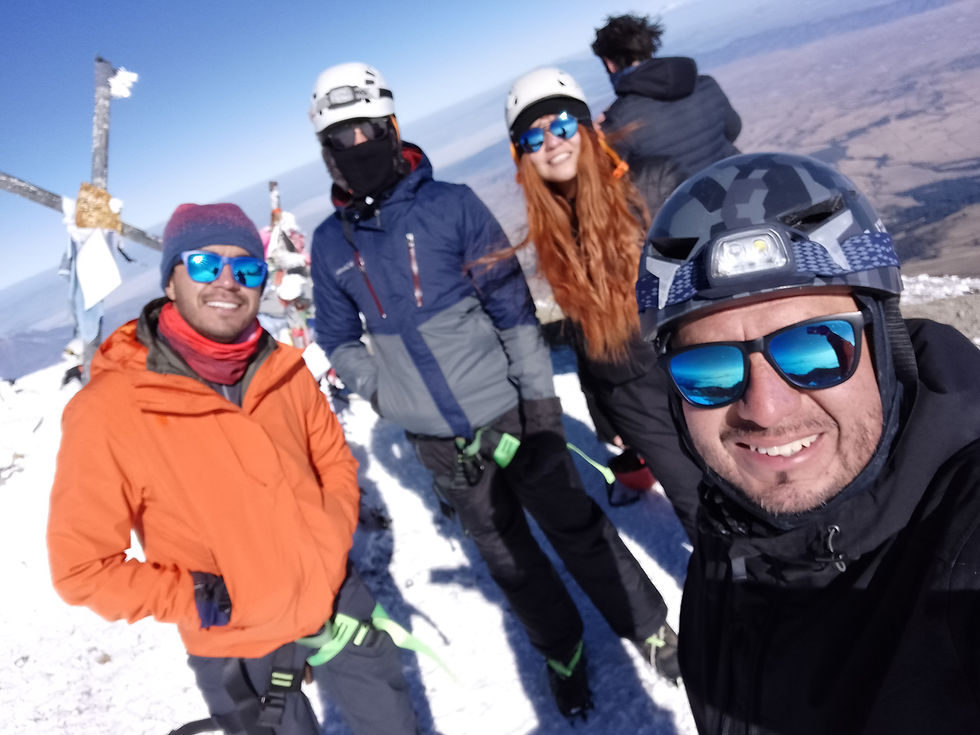 Selfie de cuatro amigos con cascos y gafas de sol en la cima del Pico de Orizaba, sonriendo con el paisaje montañoso al fondo.