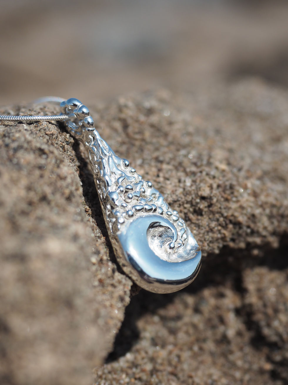 Thumbnail: Side view of teardrop shaped ocean wave silver necklace resting on sandy log
