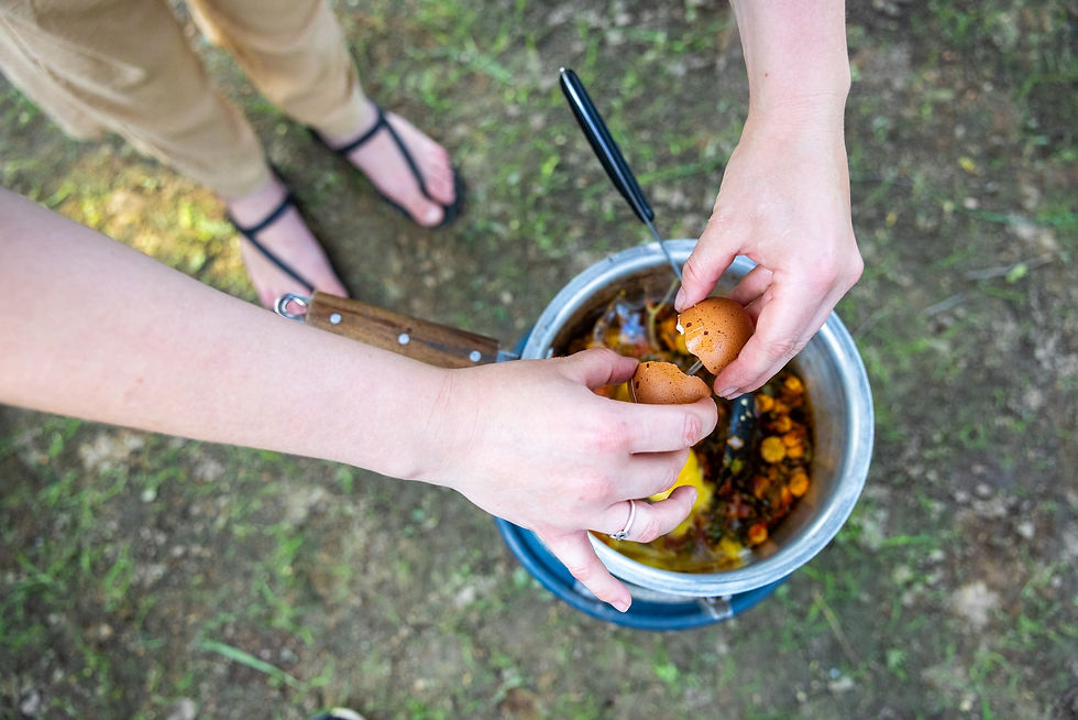 Hands crack eggs into a pot outdoors, with sandals on grass and a wooden spoon in a blue-rimmed bowl.