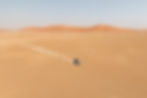 White SUV drives across a vast sandy desert, kicking up dust, with orange dunes under a pale blue sky.