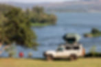 White off-road camper with rooftop tent beside two chairs overlooks a calm lake and hills on a sunny day.