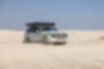 White Toyota SUV driving through sand dunes, kicking up dust, with roof rack under a clear blue sky.