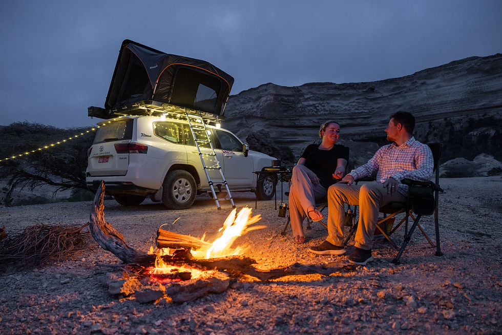 Couple sits by a campfire beside a white SUV with rooftop tent and string lights in a rocky desert at dusk.
