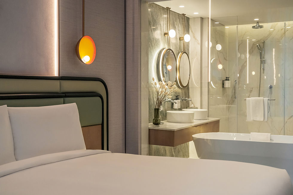 Modern bedroom with white bed beside a glass-walled marble bathroom, lit by warm lights and a glowing orange wall lamp.