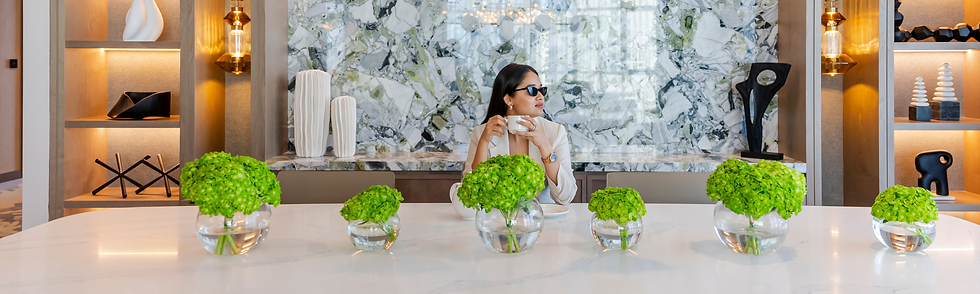 Woman in sunglasses holds a cup at a marble table with green flowers in a modern lounge with warm lit shelves.