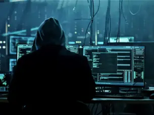 The back of a hooded individual, stealing data from computers in a dark office