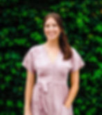Young woman with shoulder-length, brunette hair. She's wearing a light pink dress, blue, dangling earrings, and has one hand in her pocket.