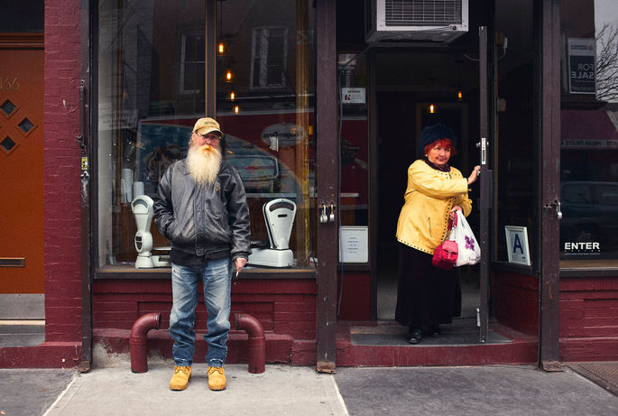 Streetphotography un homme barbu pose devant une boutique pendant qu'une femme au cheveux oranges en sort