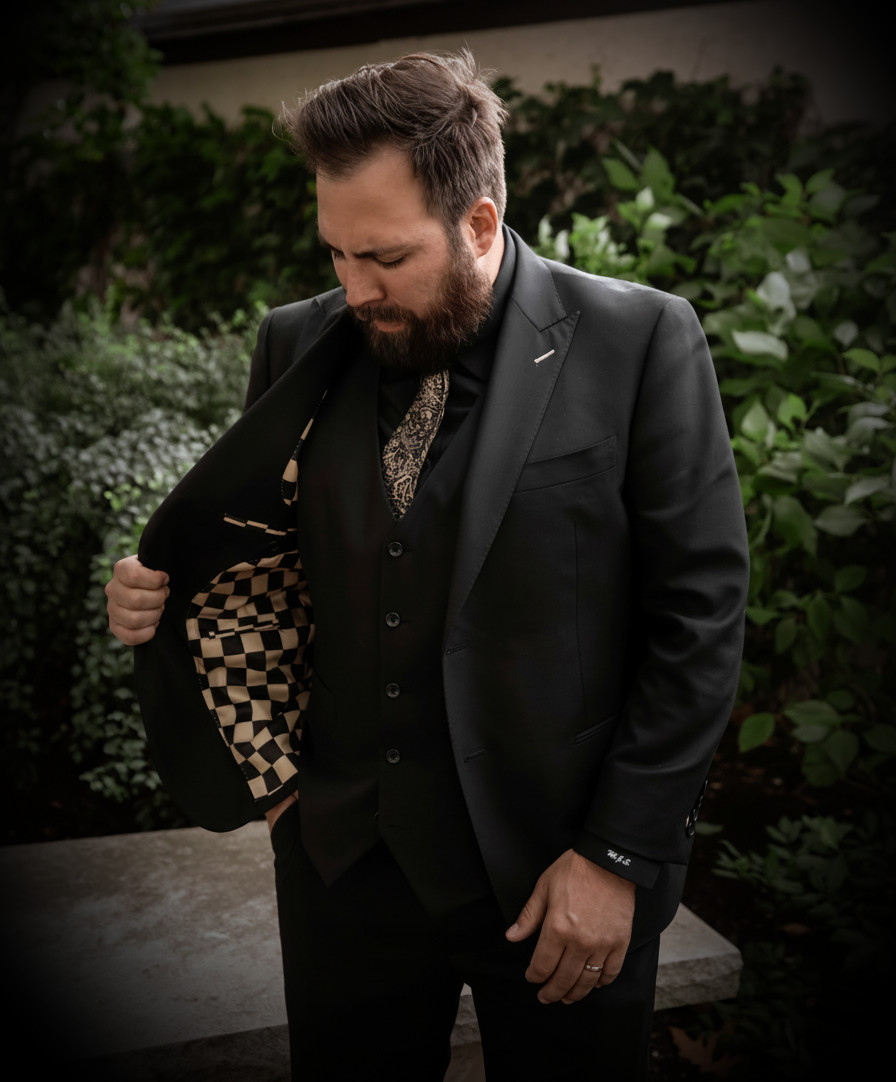 Portrait of a bearded man in a black custom suit with a patterned lining, standing outdoors with greenery in the background. Dark, cinematic, masculine vibe, portrait orientation.
