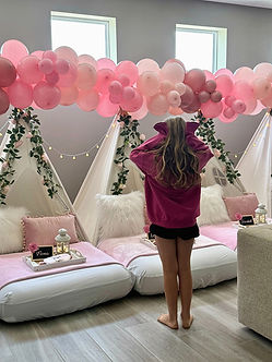 Child admiring a pink teepee sleepover party setup with styled tents, cozy bedding, and balloon garland in a bright bedroom