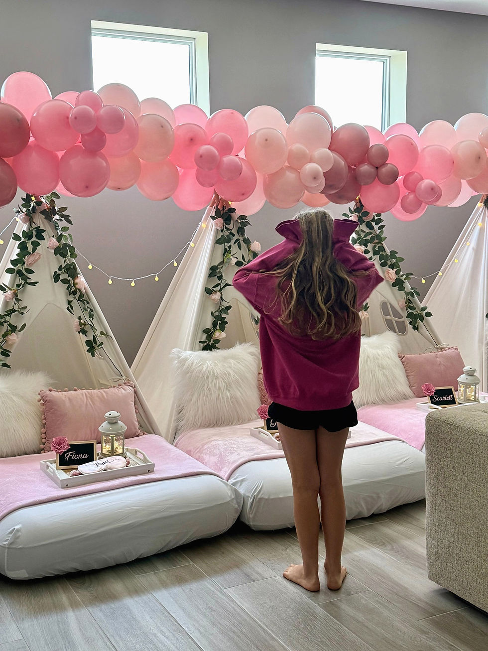 Girl in pink sweater faces cozy indoor setup with white teepees, pink bedding, and strings of lights. Pink balloons decorate above.