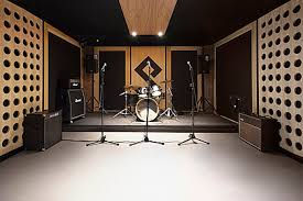 Rehearsal room with a drum set, three microphones on stands, and amplifiers. Black and wooden walls create a modern, professional vibe.