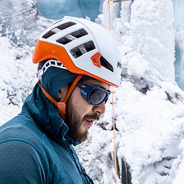 Side shot of a man wearing a helmet and hoody standing in front of an ice wall. 