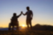 Silhouette of joyful disabled man in wheelchair raised hands with friend at sunset_edited.