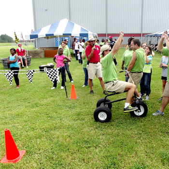 Employees trike racing at a Picnic Master company picnic, with a man on a trike high‑fiving his teammates at the finish line.