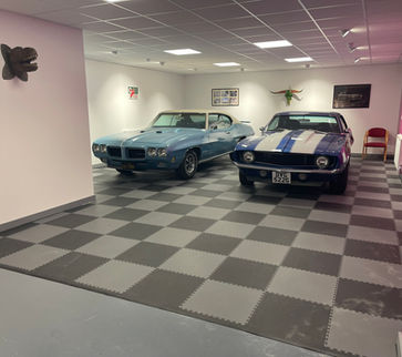 white walled room, with one fuchsia wall and chequered dark and light grey flooring, with artwork on walls and 2 vintage sports cars in the centre of the room