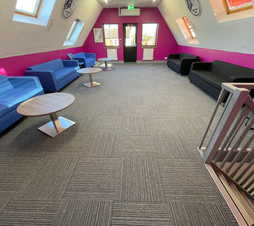 Refurbished office space with two sloped white walls, fuchsia coloured middle wall with border along the straight bottom section of the sloped walls, grey patterned carpet, 3 blue sofas with silver round coffee tables and 2 black sofas. everyone's gang logos on the 2 sloped walls