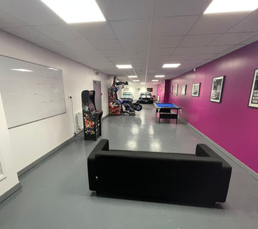 Refurbished long corridor with one white wall and one fuchsia wall, grey flooring a black sofa, several arcade games and a pool table