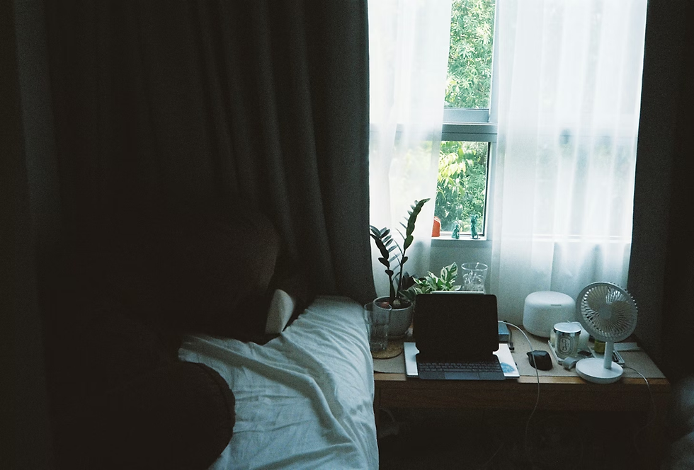 Cozy bedroom workspace with a laptop, fan, plants, and bed by a bright window with sheer and blackout curtains and green trees outside.
