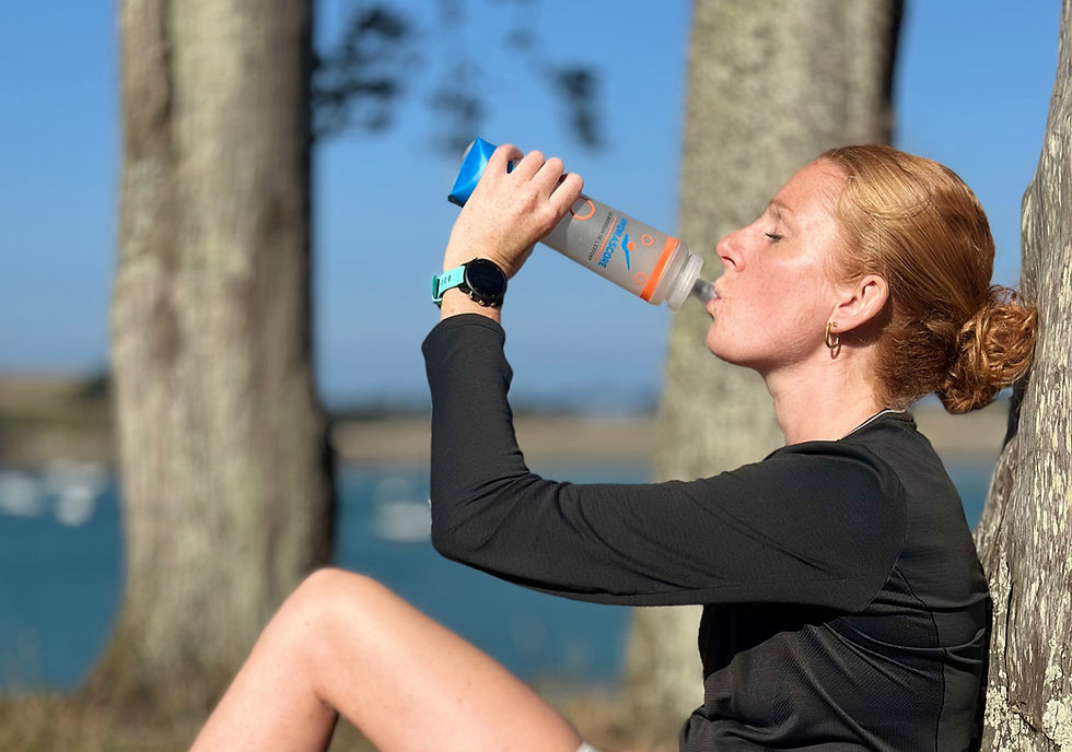 Femme boit de l'eau d'une flasque Hydrascore, assise devant des arbres.