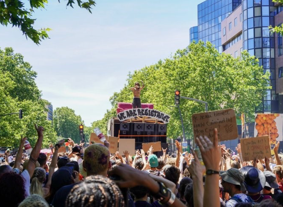 Une foule de participant·es danse lors d'une free party en plein air, devant un mur d'enceintes et sous des jeux de lumière colorés. L'image illustre la culture des teufs et les rassemblements musicaux alternatifs souvent visés par les politiques de maintien de l'ordre. - Bloc Populaire