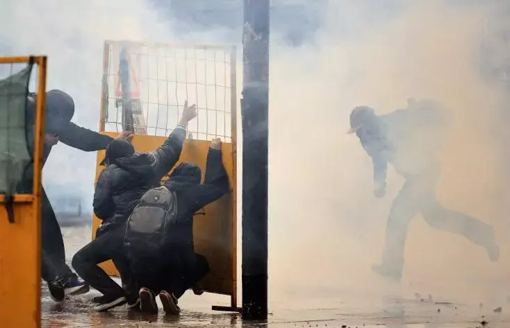 Manifestants accroupis derrière une barrière, dans le gaz lacrymogène, tandis qu’un homme fuit à droite.