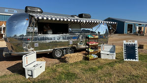 Silver Airstream food trailer labeled "Artisan & Vine Market" with striped awning, surrounded by hay bales, snacks, coolers, and a menu sign, under clear skies.