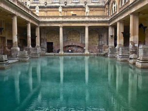 A wide stone pool surrounded by Roman columns, known as the Great Bath in Bath, England, filled with turquoise water from natural hot springs.