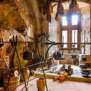 Authentic medieval kitchen with stone walls, hanging meats, dried herbs, iron pots, and clay vessels, showing how food was stored and cooked in daily life.