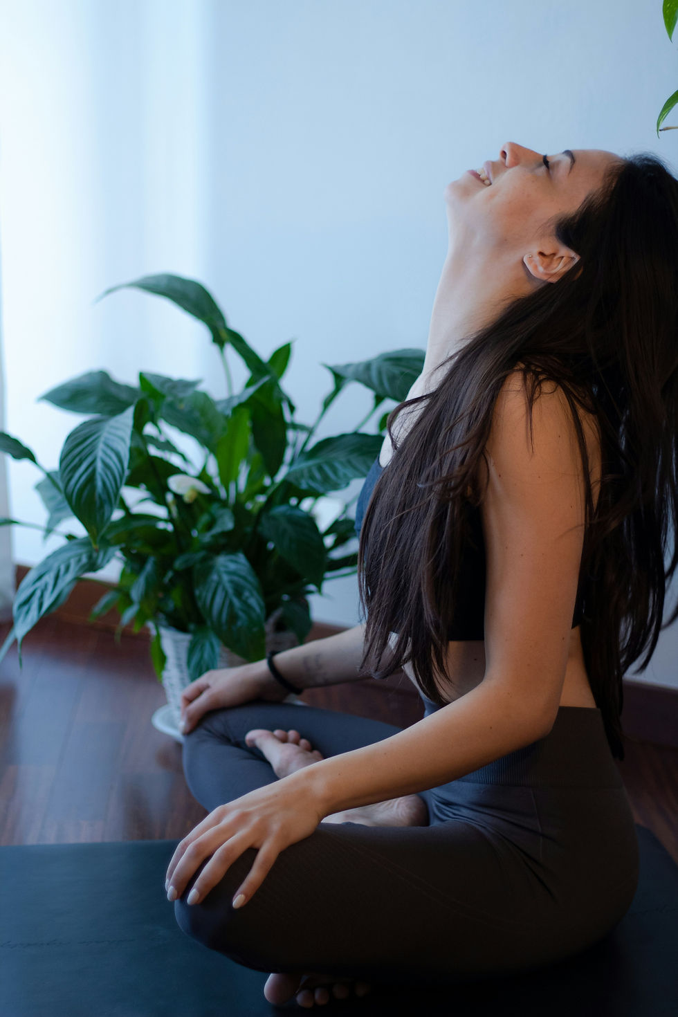 Woman in yoga pose, sitting cross-legged on a mat, head tilted back, smiling. Green plant in the background, wooden floor visible.