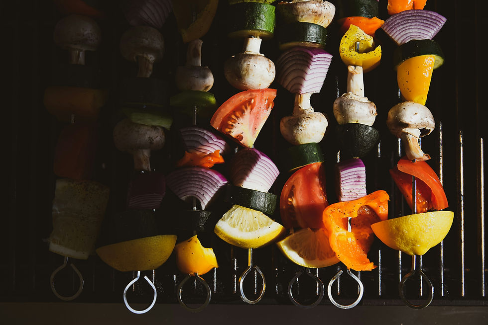 Vegetable skewers on a grill with mushrooms, tomatoes, onions, peppers, and lemon slices. Sunlight casts shadows and highlights vibrant colors.