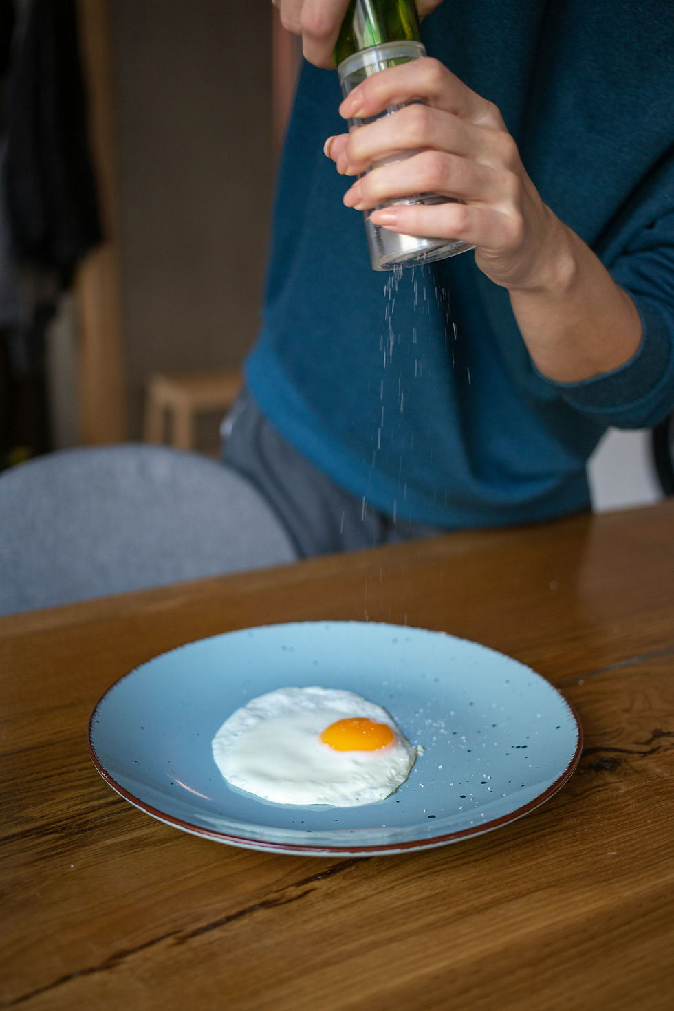 A person sprinkles salt on a fried egg on a blue plate. The plate is on a wooden table.