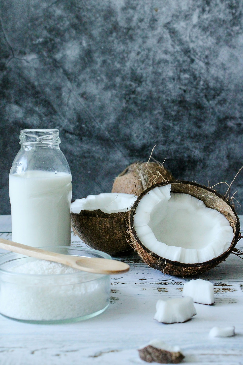 Coconut halves, a bottle of coconut milk, and grated coconut in a bowl with wooden spoon on a white surface with a textured gray background.