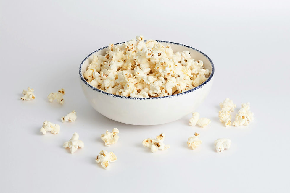 A white bowl filled with fluffy popcorn sits on a white surface. A few kernels are scattered around. The mood is light and casual.