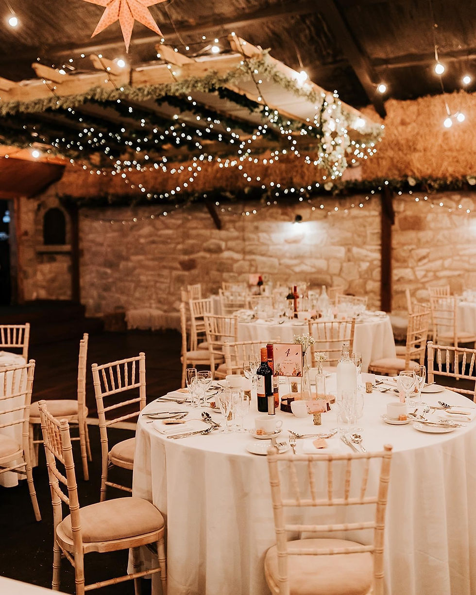 Elegant barn with round tables set for a formal dinner, warm lighting, rustic stone walls, and twinkling fairy lights overhead.