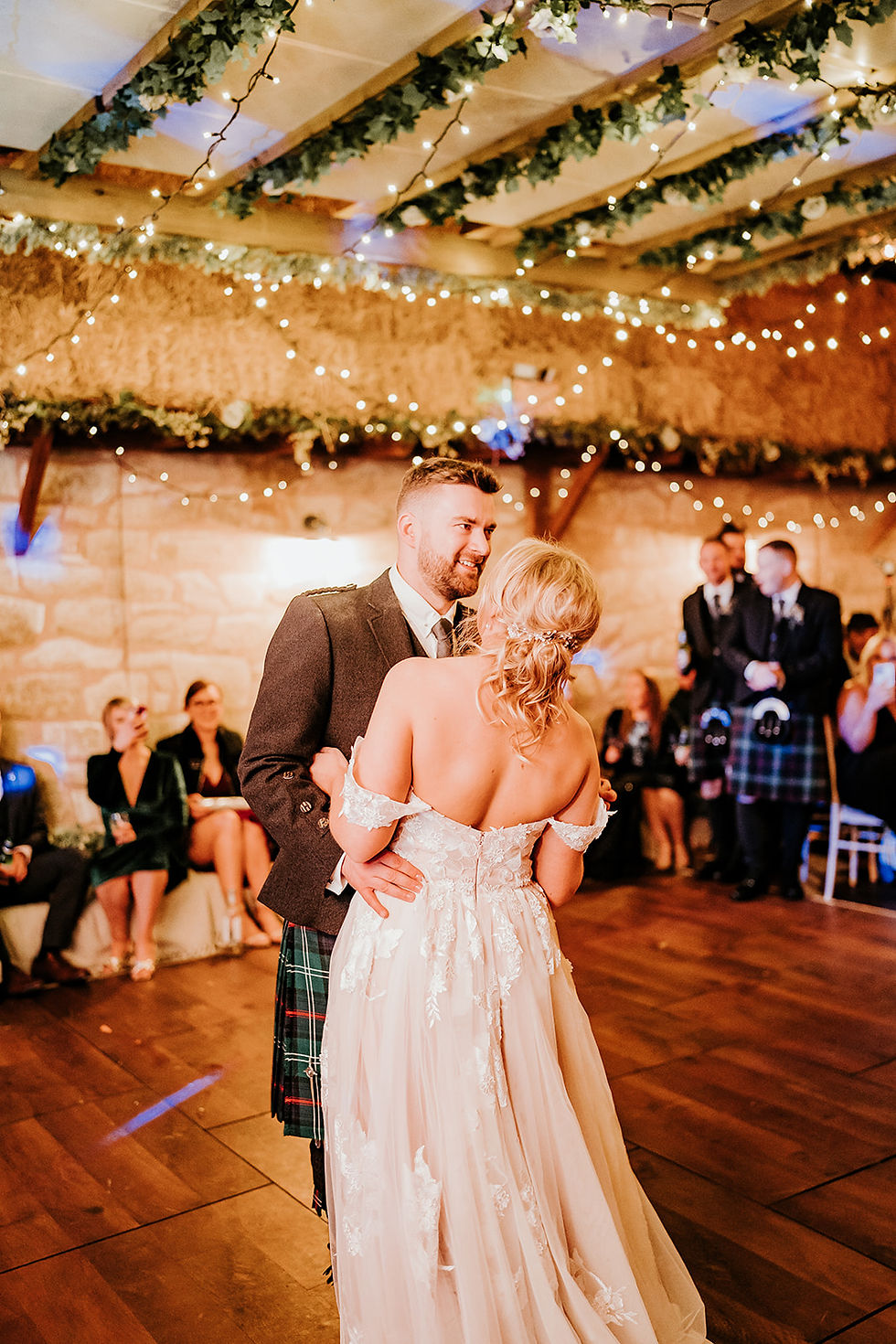A couple dances under fairy lights in a rustic venue. The woman wears a white gown; the man in a kilt smiles. Guests are watching.