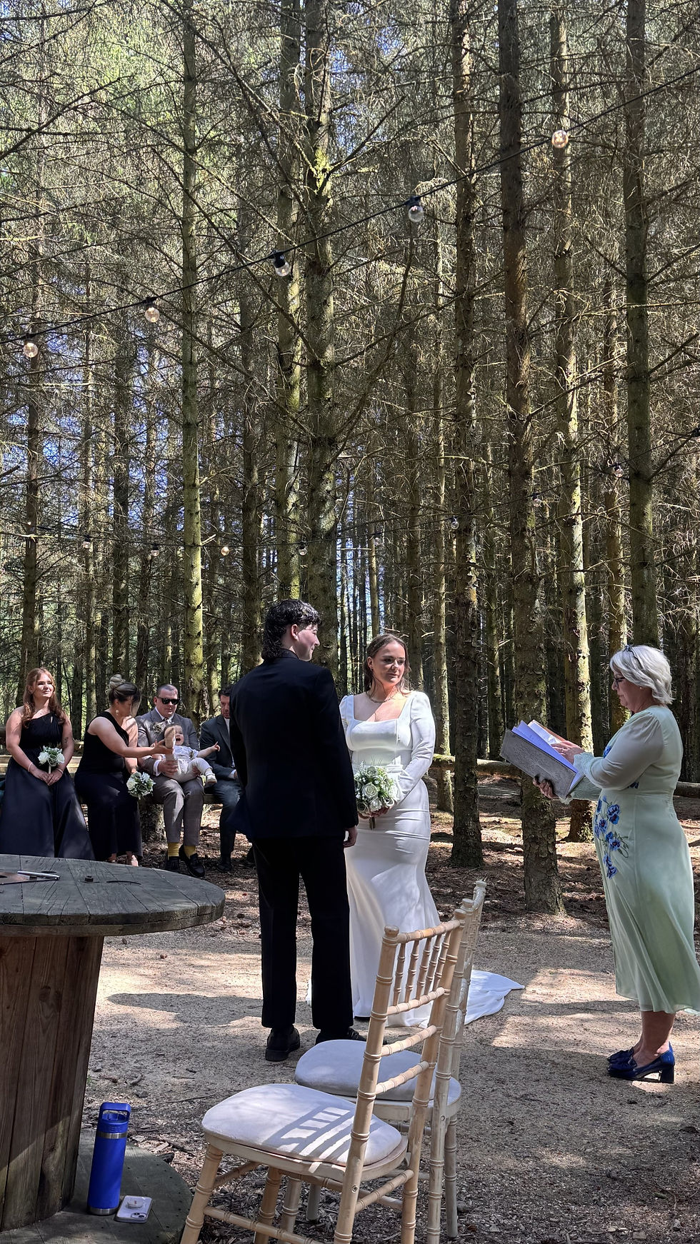 Bride and groom stand at a forest wedding ceremony while an officiant reads, guests seated under string lights.