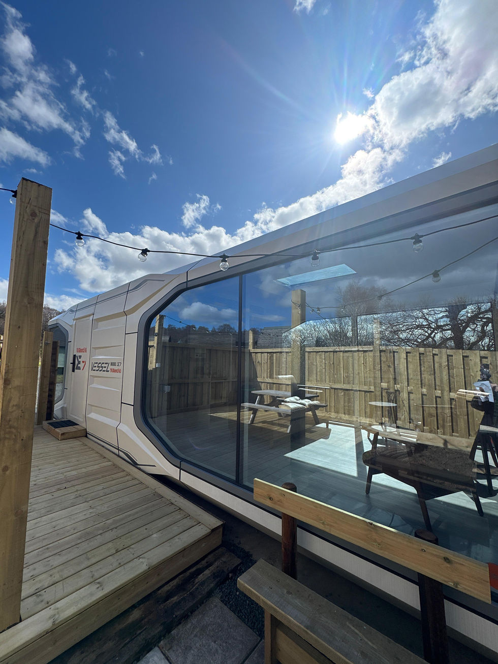 Sunlit modern white VESSEL pod with large glass wall on a wooden deck, string lights overhead, blue sky and clouds reflected in glass.