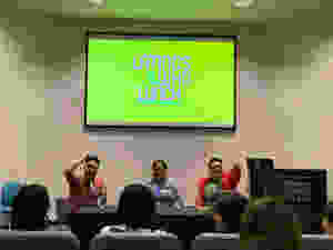 Emmanuel Ortega, Erika Abad, and Justin Favela sit at a table facing a crowd of listeners while smiling and gesturing to the audience. The Latinos Who Lunch logo is projected on the screen behind them in vivid lime green.