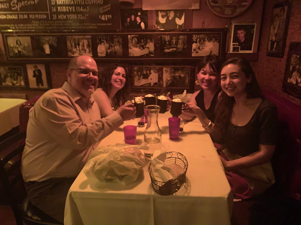 Jacob, Alison, Kristy & Sonja sit at a white tablecloth dinner holding mugs of hot chocolate with whipped cream. The lighting is dim and there are rows of black and white photographs filling the wall behind them.