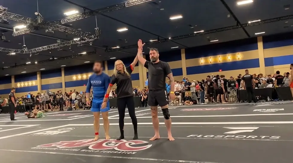 Event photography featuring a referee raising the arm of a victorious martial arts competitor at a tournament. The scene captures a triumphant moment on the mat with spectators visible in the background.