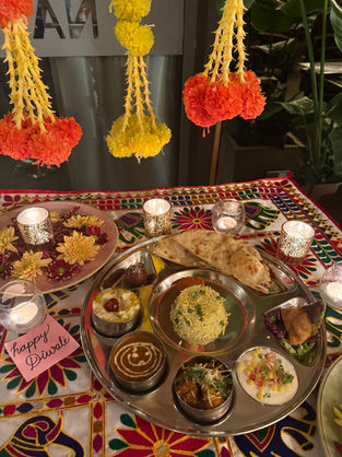 Traditional Indian meal on a vibrant tablecloth with marigold garlands above. A note reads "happy Diwali." Candles and flowers add warmth.