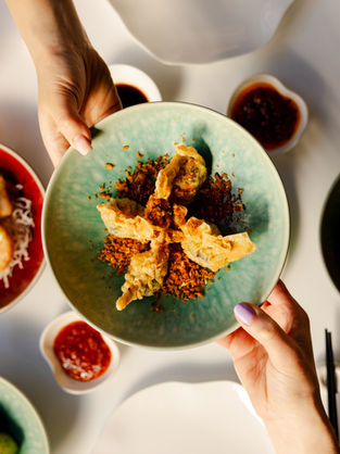 Two hands holding a green plate with fried dumplings and crispy bits. Background features dipping sauces and dishes on a light table.