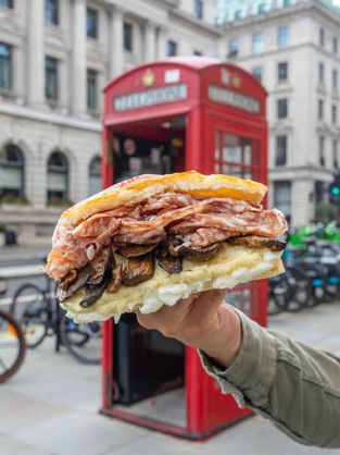 A hand holding a large sandwich filled with meat and mushrooms. A red British phone booth and bicycles are in the city background.