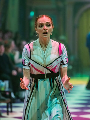 Performer in a colorful dress singing passionately on a checkered stage with a blurred audience and dancers in the background.