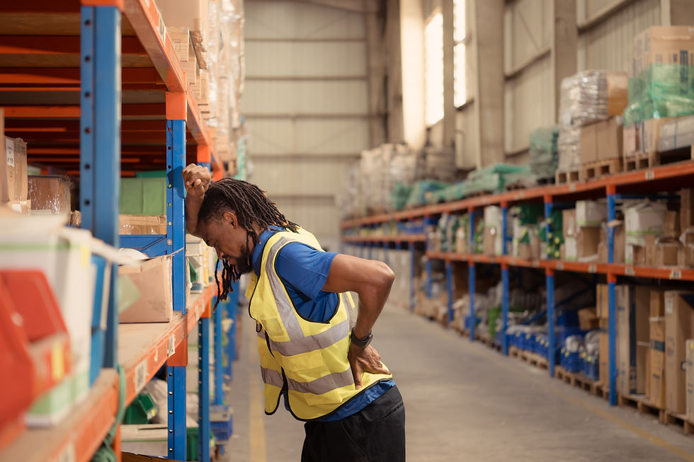 Worker in a yellow vest leans on a shelf, hand on back, in a warehouse aisle filled with boxes, appearing injured.