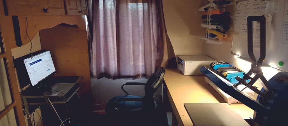 Small home office with desk, monitor, office chair, purple curtains, whiteboard and fabric rolls, warmly lit and tidy.
